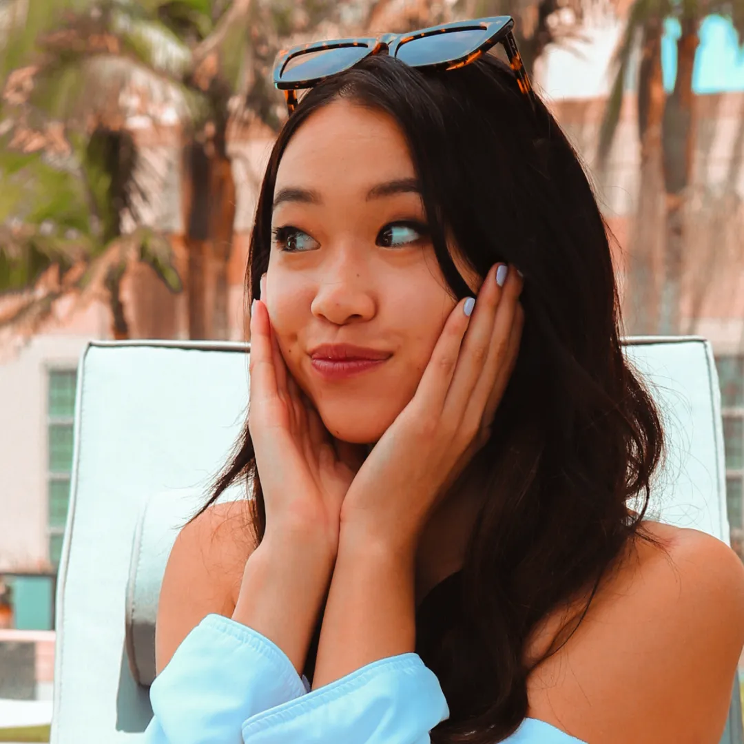 A woman in a light blue top smiles playfully with sunglasses on her head, in warm outdoor lighting.